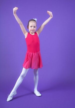 Roter Tanzanzug mit einem ausgestellten rosa Rock, weißen Strumpfhosen und weichen weißen Ballettschuhen. Der Hintergrund ist uniform lila. Die Hände sind in einer Pose erhoben.