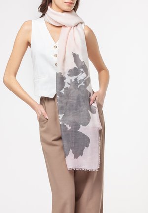 Donna che indossa una sciarpa rosa chiaro con grande stampa floreale grigia, gilet bianco senza maniche con bottoni e pantaloni beige a vita alta.
