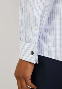 Camisa de manga larga blanca con un patrón de cuadros azules, puños de botón y un botón decorativo con detalle del logotipo. Apta para vestimenta formal.
