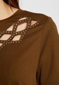 Brązowa bluzka z diamentowymi wycięciami, ozdobiona małymi srebrnymi ćwiekami. Gładka w fakturze, z okrągłym dekoltem i krótkimi rękawami.