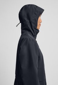 Schwarze Kapuzenjacke, strukturiertem Stoff, elastischem Kordelzug an der Kapuze, taillierter Schnitt, seitlichen Taschen und einer leichten Raffung an der Taille.