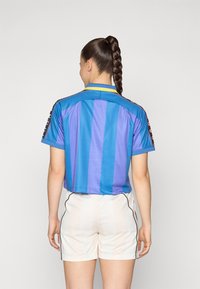 Kék és lila csíkos sportmez sárga gallérral és fekete díszítésekkel, fehér sorttal, oldalcsíkokkal párosítva.