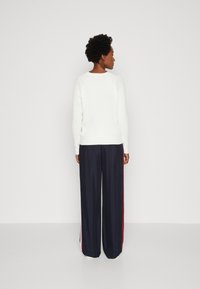 Maglione bianco a coste con scollo rotondo, abbinato a pantaloni larghi blu navy con strisce rosse sui lati. Vista posteriore.
