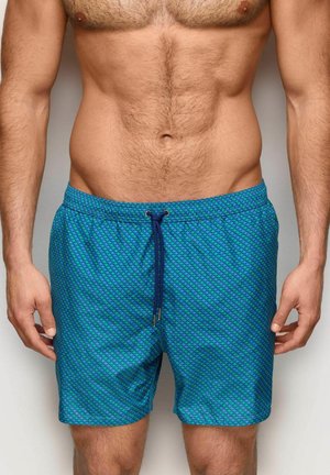 Torso di un uomo senza maglietta che indossa pantaloncini da bagno blu a fantasia con coulisse blu scuro, in piedi con le braccia rilassate lungo i fianchi.