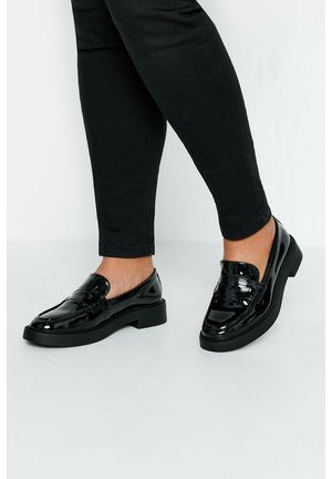 Mocasines de charol negro con puntera redondeada, detalles texturizados en el empeine y un tacón bajo de bloque. Llevados con pantalones ajustados negros.