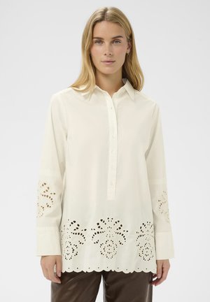 Szőke nő, aki fehér, hosszú ujjú blúzt visel csipkés kivágásokkal az ujjakon és az alsó részen, sötétbarna nadrággal párosítva.