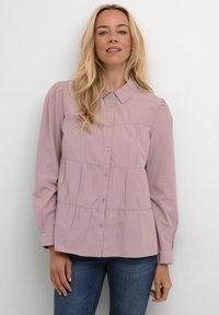 Camicia botton-down di colore lilla chiaro con colletto, caratterizzata da un design a strati e maniche lunghe. Tessuto liscio con sottili dettagli arricciati.