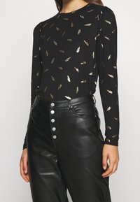 Schwarzes Langarmshirt mit goldenen Feder-ähnlichen Mustern, aus weichem Material, kombiniert mit hochgeschnittenen schwarzen Lederhosen mit silbernen Knöpfen.