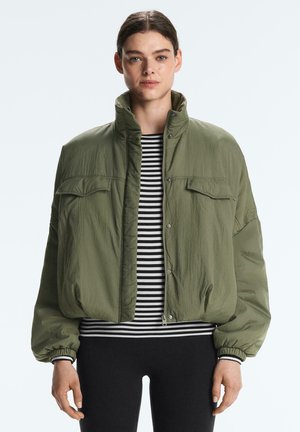 Frau trägt locker sitzende olivgrüne Jacke mit Klappentaschen über schwarz-weiß gestreiftem Shirt und schwarzer Hose vor neutralem Hintergrund.