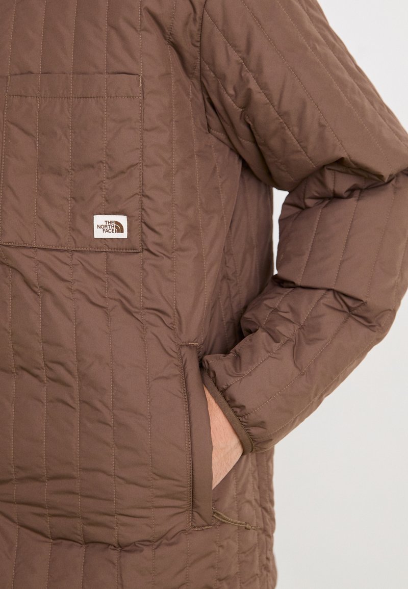 Giacca imbottita marrone con tasca sul petto e aperture laterali. Realizzata in tessuto leggero con una texture liscia e una leggera lucentezza.