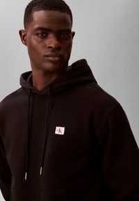 Mladić u crnoj kapuljači s logotipom Calvin Klein na prsima, okrenut prema naprijed na jednobojnoj sivoj pozadini.