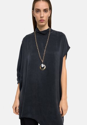 Kobieta ubrana w luźną, ciemnoszarą tunikę z wysokim kołnierzem oraz długi łańcuch z dużymi, okrągłymi srebrno-złotymi zawieszkami.