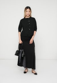 Black cardigan with button closure over a black maxi skirt. Slim-fit design, smooth texture. Accessorized with a black handbag and polka dot scarf.