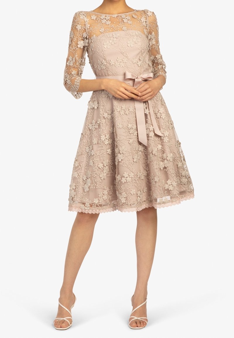 Robe beige mi-longue avec manches 3/4 en dentelle florale transparente, broderies florales et nœud en satin à la taille, associée à des escarpins blancs à brides.