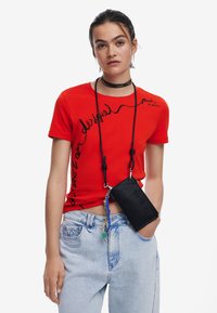 Czerwona koszulka z krótkim rękawem z czarnym kursywnym napisem, zestawiona z jasnoniebieskimi dżinsami. Czarna saszetka przyczepiona paskami z kolorowymi zawieszkami.