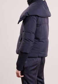 Chaqueta de plumas azul marino con un único cuello sobredimensionado, diseño acolchado, puños acanalados y un corte ajustado, combinada con pantalones a rayas.