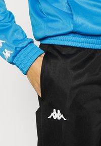 Ruka v čiernych nohaviciach, oblečený v modrej bunde s bielymi logami na rukáve a nohaviciach na jednofarebnom pozadí.