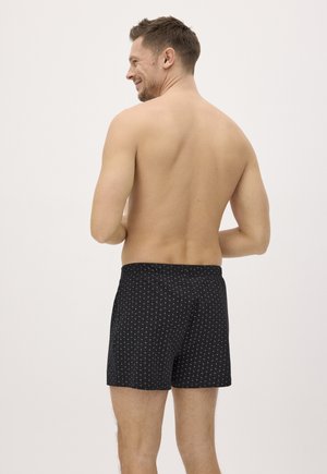 Hombre de espaldas vestido con bóxers negros con estampado, sonriendo, con torso desnudo sobre un fondo claro y liso.