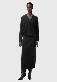 Svart stickad cardigan med V-ringning och knappar, ihopparad med en lång svart kjol. Outfitten har en texturerad finish och minimalistisk design.