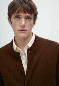Brauner gestrickter Cardigan mit hohem Rippenkragen, ausgestattet mit zwei schwarzen Knöpfen und getragen über einem weißen Hemd mit Kragen. Glatte Textur.