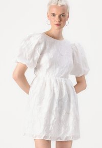 Λευκό φόρεμα με φουσκωτά μανίκια, κατασκευασμένο από υφασμάτινη ύφανση με λουλούδια, με στενή μέση και κοντό μήκος. Διαθέτει στρογγυλή λαιμόκοψη και παιχνιδιάρικη σιλουέτα.