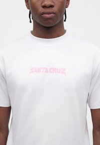 Νεαρό άτομο με κοτσίδες που φοράει άσπρο T-shirt με μικρό ροζ λογότυπο «Santa Cruz» στο στήθος.