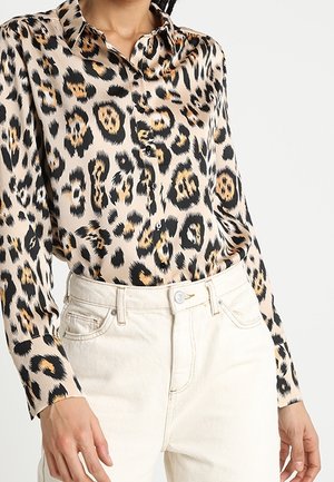 Vrouw draagt een beige blouse met lange mouwen en luipaardprint, ingestopt in een hooggetailleerde off-white jeans met voorzakken en knoopsluiting.