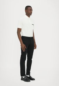 Cămașă polo albă cu un buzunar negru, asortată cu blugi negri și mocasini negri. Ținuta are un design ajustat și texturi fine.