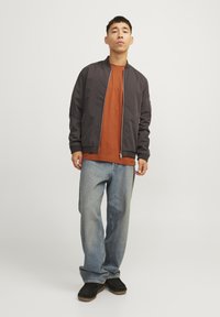 Lightweight black bomber jacket over an orange shirt, paired with loose blue jeans and black slip-on shoes. Simple design with a zip closure.