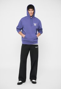 Felpa viola con cappuccio e tasca frontale, con logo bianco. Abbinata a pantaloni neri a gamba larga e scarpe nere. Design semplice e vestibilità casual.