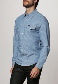 Vaaleansininen farkkupaita, pitkät hihat, jämäkkä kaulus, kaksi rintataskua ja napillinen edusta. Pehmeä rakenne ja klassinen istuvuus.