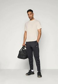 Mies, jolla on päällään beigepaita ja mustat Nike-housut, joka pitää mustaa urheilukassia kädessään ja seisoo yksinkertaista vaaleaa taustaa vasten.