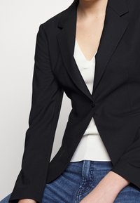 Černý upravený blazer s jedním knoflíkem, strukturovanými rameny a klopami, vrstvený přes bílý žebrovaný top. Viditelné džíny.