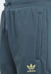 Gröna joggers i mjukt material, med snörning i midjan, sidofickor och en gyllene Adidas-logga på vänster lår.