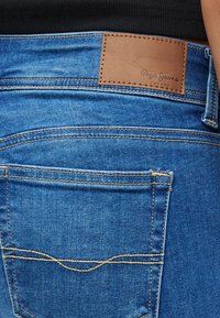 Kék farmernadrág barna bőrpöttyel, rajta dombornyomott logóval. Látható varrásrészlet és klasszikus zsebminta a hátsó részen.