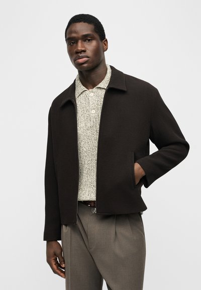 Νέος άντρας που φοράει μαύρο μπουφάν πάνω από ένα ανοιχτόχρωμο πουκάμισο με υφή και κουμπιά, καθώς και γκρι πλισέ παντελόνια, με το ένα χέρι στην τσέπη του μπουφάν.