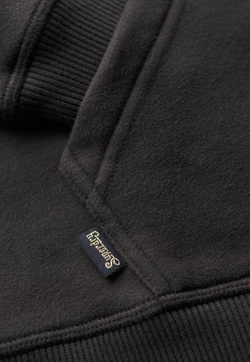 Schwarzer Pullover mit gerippten Bündchen, weicher Textur und einem kleinen, goldgestickten Etikett mit der Aufschrift "Superdry" auf einem navy Hintergrund.