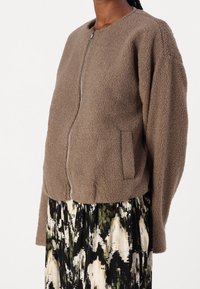 Osoba v hnědé fleecové bundě na zip s bočními kapsami, pod kterou má na sobě sukni s abstraktním vzorem v černé, bílé a zelené barvě.