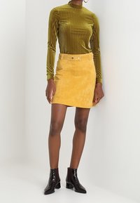 Haut à manches longues en velours côtelé vert, mini-jupe en daim moutarde avec ceinture, et bottines noires en cuir verni. Design simple, textures lisses.