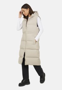 Gilet imbottito senza maniche beige con cappuccio, chiusura con zip, due tasche frontali e design lungo fino al ginocchio, abbinato a pantaloni neri e stivali.