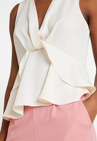 Hihaton pusero pehmeästä norsunluun värisestä kankaasta, jossa on drapeerattu etuosa kiertozepillä ja laskeutuva röyhel�öhelma, yhdistetty vaaleanpunaisten alaosien kanssa.