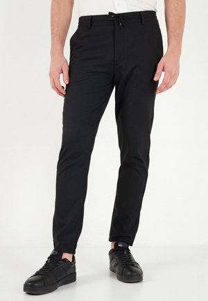 Homme portant un pantalon noir slim avec une taille à cordon et des baskets basses noires, debout sur un fond blanc.