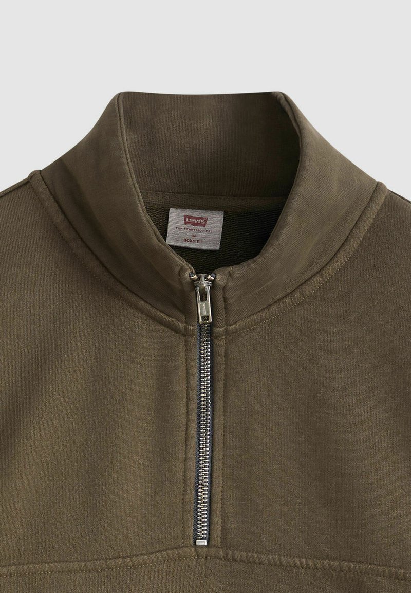 Pull-over vert olive avec un col montant et une fermeture éclair quart de zip. Confectionné dans un tissu lisse et texturé avec une coupe sportive et des détails de couture.