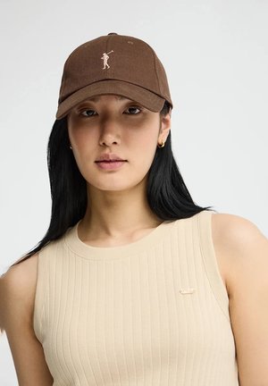 Giovane donna con lunghi capelli neri, che indossa un cappellino da baseball marrone e un top beige senza maniche a coste, rivolta verso l'obiettivo su uno sfondo uniforme.