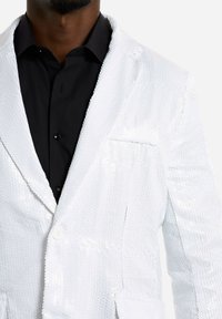 Weißer blumiger Blazer mit strukturiertem Design, Ein-Knopf-Verschluss und einer Tasche. Getragen über einem schwarzen Hemd mit Kragen.