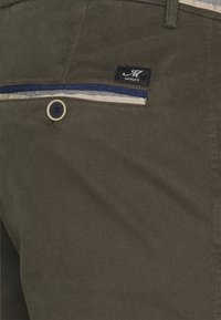 Pantaloni verde oliva realizzati in tessuto testurizzato, con chiusura a bottone e rifinitura delle tasche in blu e beige a contrasto, completa di etichetta del marchio.