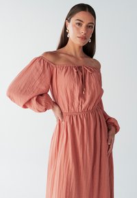 Robe corail rose à épaules dénudées avec de longues manches bouffantes, taille cintrée et tissu texturé. Présente un détail à lien au niveau du col.