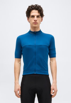 Jeune homme aux cheveux ondulés portant un maillot de cyclisme bleu ajusté et un cuissard rembourré noir, debout devant un fond uni.