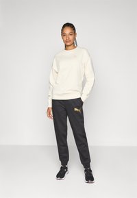 Cremefarbener Pullover mit kleinem Logo, kombiniert mit dunklen Hosen, die ein goldenes Logo haben. Schwarze Sportschuhe runden den Look ab.