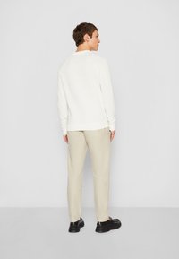 Weißer strukturierter Pullover mit langen Ärmeln, beiger Hose und schwarzen Schuhen. Die Ansicht ist von hinten und zeigt eine lockere Passform sowie ein einfaches Design.